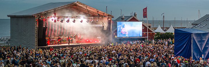 En upplyst artiscen med ett folkhav framför. En artist uppträder på scenen. Foto.